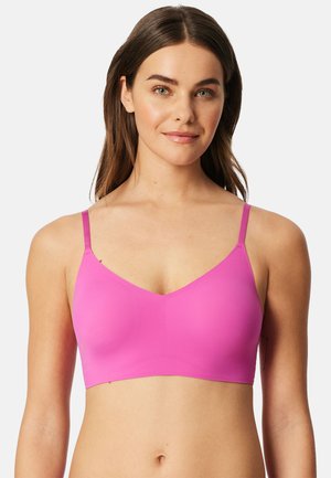 Joven mujer con cabello castaño liso, vistiendo un sujetador sin costuras de color rosa brillante con tirantes delgados, sonriendo suavemente frente a un fondo liso.