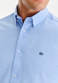 Jasnoniebieska koszula z kołnierzykiem typu button-down, z białymi guzikami i czarnym logo "PJ" na obszarze klatki piersiowej.