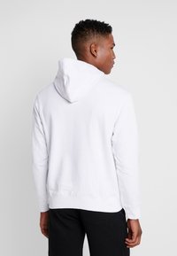 Sudadera blanca con capucha, de manga larga, con un ajuste relajado, puños y bajo acanalados. Vista trasera, la tela parece suave y ligeramente texturizada.