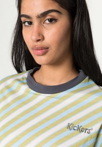 T-shirt às riscas em azul claro, verde e branco. Apresenta um colarinho redondo em azul-marinho e o logótipo bordado "Kickers" em cinza. Material de algodão suave.