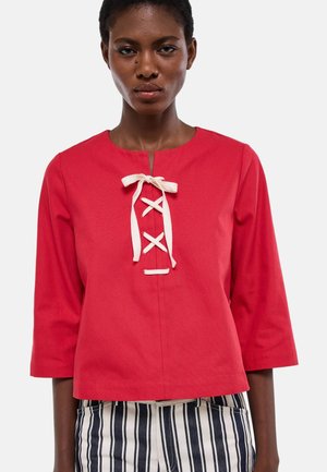 Femme portant un haut rouge à manches trois-quarts avec un détail lacé blanc au niveau de l'encolure et un pantalon à rayures verticales noires et blanches.