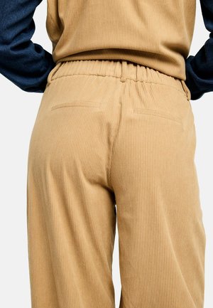 Person in beigen Cordhosen und passendem Oberteil mit elastischem Bund und Gesäßtaschen, steht mit den Händen in der Hüfte.