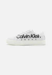 Vit låg sneakers med svart "Calvin Klein Jeans"-logga på sidan och präglad branding på sulan mot en enfärgad bakgrund.