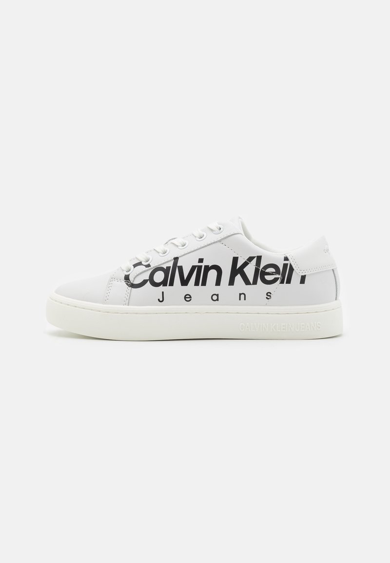Vit låg sneakers med svart "Calvin Klein Jeans"-logga på sidan och präglad branding på sulan mot en enfärgad bakgrund.