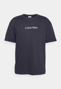 Námořnická modrá bavlněná tričko s krátkým rukávem, kulatým výstřihem a bílým logem "Calvin Klein" vytištěným na hrudi. Měkká textura, uvolněný střih.
