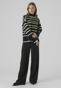 Pull rayé noir et crème avec un col montant et des manches larges, porté avec un pantalon noir ample et des chaussures noires à accents floraux.