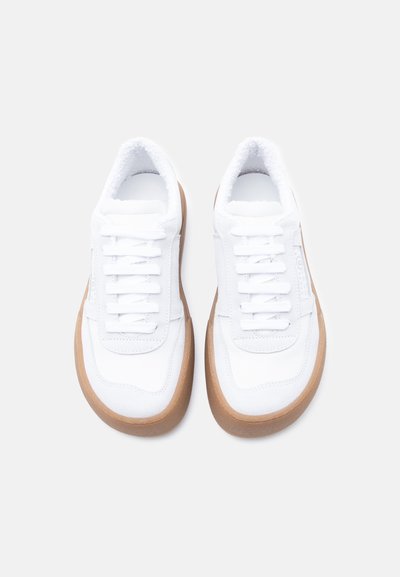 Baskets blanches avec tissu texturé, lacets plats, col rembourré et semelle en caoutchouc marron, avec un logo subtil sur le côté.
