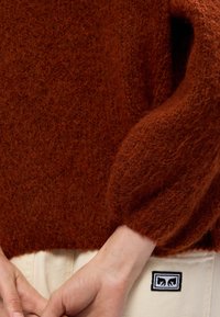 Rustfarvet sweater lavet af blødt, struktureret garn, med en afslappet pasform og samlede ærmer. Inkluderer et lille mærke på kanten.