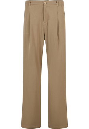 Homme debout, les mains dans les poches, portant une chemise blanche à manches courtes oversize, un pantalon beige ample et des baskets blanches sur fond gris uni.