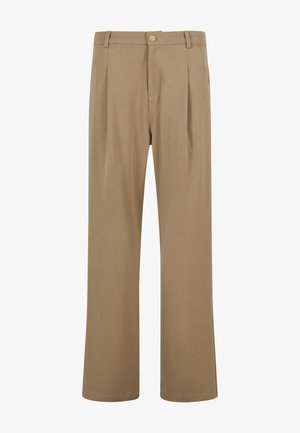 Homme debout, les mains dans les poches, portant une chemise blanche à manches courtes oversize, un pantalon beige ample et des baskets blanches sur fond gris uni.