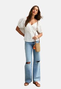 Blouse blanche à manches bouffantes avec des détails en dentelle et fermeture à boutons, associée à un jean flare taille haute bleu clair avec des déchirures aux genoux. Sandales marron et sac en osier.
