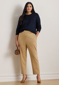 Moteris apsivilkusi tamsiai mėlyną megztinį, smėlio spalvos aukštai supintas kelnes, rudas aukštakulnius ir mažą rudą rankinę, stovinti prieš paprastą šviesią sieną.