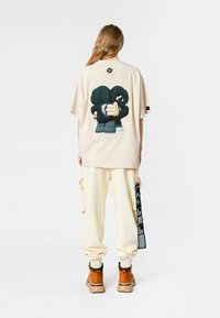 Camiseta de gran tamaño en color beige con una gráfica de dos figuras abstractas abrazándose en la parte posterior. Combinada con pantalones cargo color crema y calzado marrón.