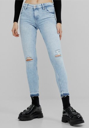 Jasnoniebieskie podarte wąskie jeansy noszone z czarnymi skarpetkami i masywnymi czarnymi butami, stylizowane z krótkim czarnym topem z długim rękawem.