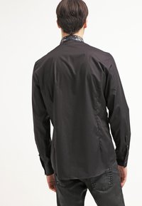 Camicia nera a maniche lunghe con una texture liscia e un colletto decorato. Presenta polsini con bottoni e un orlo leggermente curvato.