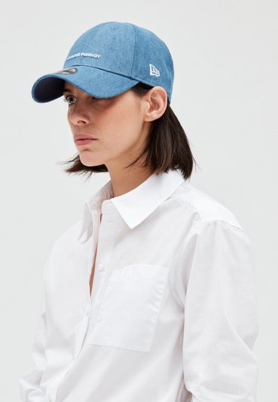 Denim kék baseball sapka ívelt csúccsal és hímzett logóval. Fehér galléros inggel viselve. Egyszerű és díszítetlen design.