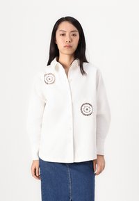Cămașă albă cu guler clasic, mâneci lungi și accente de dantelă circulară brodată pe piept, asociată cu o fustă din denim.