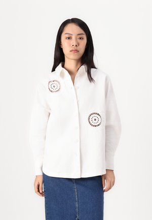 White button-up shirt with embroidered circular lace accents on the chest, long sleeves, and a classic collar, paired with a denim skirt.