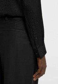 Camicia nera a maniche lunghe con un motivo testurizzato, abbinata a pantaloni neri su misura. La camicia presenta polsini con bottoni e dettagli tonali.
