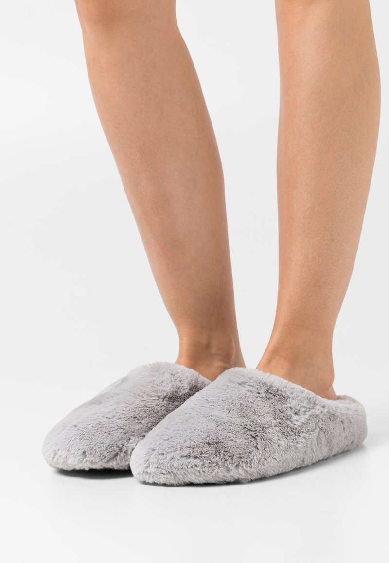 Paire de jambes nues portant des chaussons légers, gris clair et doux, sur un fond blanc.