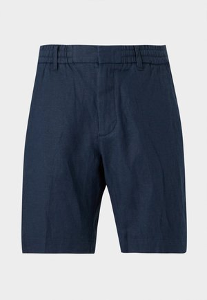 Mann steht mit Händen in den Taschen, trägt ein beiges kurzärmeliges Hemd mit Knöpfen, marineblaue Shorts und schwarze Sandalen vor einem schlichten Hintergrund.