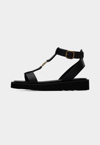 Μαύρο δερμάτινο σανδάλι με σχέδιο T-strap, ρυθμιζόμενο πόρπη στον αστράγαλο και παχιά, υφή σόλα. Διαθέτει χρυσούς μεταλλικούς τόνος διακοσμητικά στοιχεία.