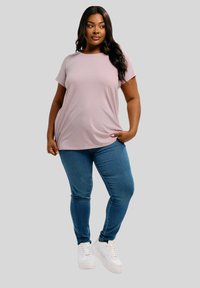 Donna con lunghi capelli scuri che indossa una maglietta rosa chiaro, jeans blu e sneakers bianche, in piedi con una mano nella tasca su uno sfondo grigio.