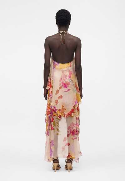 Γυναίκα με κοντά μαλλιά, ντυμένη με διαφανές, λουλουδάτο, αμάνικο φόρεμα με ανοιχτή πλάτη και μπεζ πέδιλα με τακούνι, στέκεται με φόντο απλό λευκό.