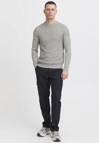 Grey ribbed knit sweater, dark cargo pants, and grey athletic shoes. Model stands with hands in pockets against a plain background.