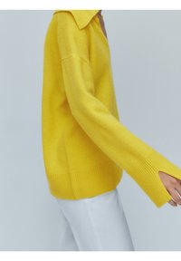 Geel gebreid sweater met een kraag, brede mouwen met sleuven en een geribbelde zoom. Gecombineerd met witte broek, tegen een neutrale achtergrond.