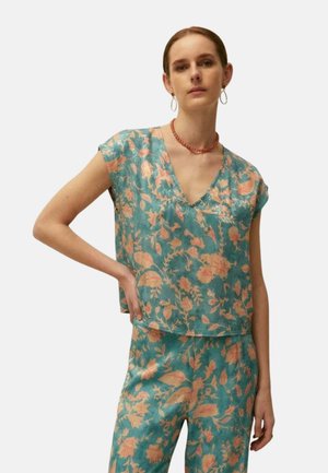 Mujer con camiseta de manga corta floral en color aguamarina y melocotón y pantalones a juego, posando con una mano en la cadera sobre un fondo liso.