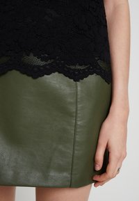 Haut en dentelle noire à motif floral superposé à une jupe en cuir verte. La jupe présente une texture lisse et un détail de couture latérale.