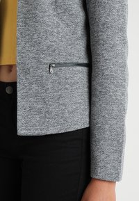 Šedá texturovaná bunda s elegantním designem a boční kapsou na zip. Materiál vypadá měkce a pleteně, v kombinaci s kontrastním žlutým topem.