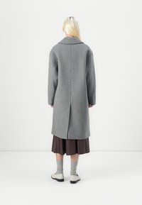 Lungo cappotto di lana grigia con colletto, vestibilità over, e spacco posteriore, abbinato a una gonna plissettata marrone e calzini grigi. Visto da dietro.