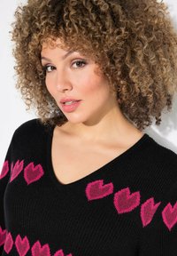 Maglione nero a maglia con scollo a V, decorato da motivi a cuore rosa sul petto. Texture morbida e spessore medio.