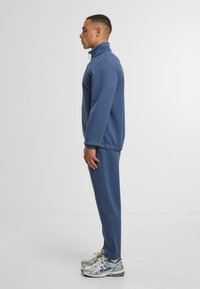 Zils flīsa apgriezts jaka ar augstu apkakli, apvienots ar atbilstošām zilām sporta biksēm. Pelēki un zili sporta apavi papildina apģērbu.