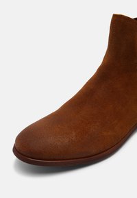 Botte cheville en daim brun avec un bout rond et un design minimaliste. La texture est douce, avec une surface lisse et une semelle en caoutchouc à profil bas.