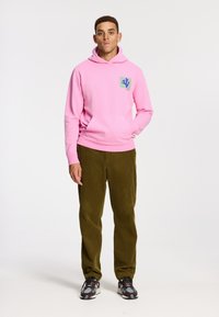 Felpa rosa con tasca frontale e un patch logo colorato sul petto, abbinata a pantaloni in corduroy verde oliva e sneakers con motivo.