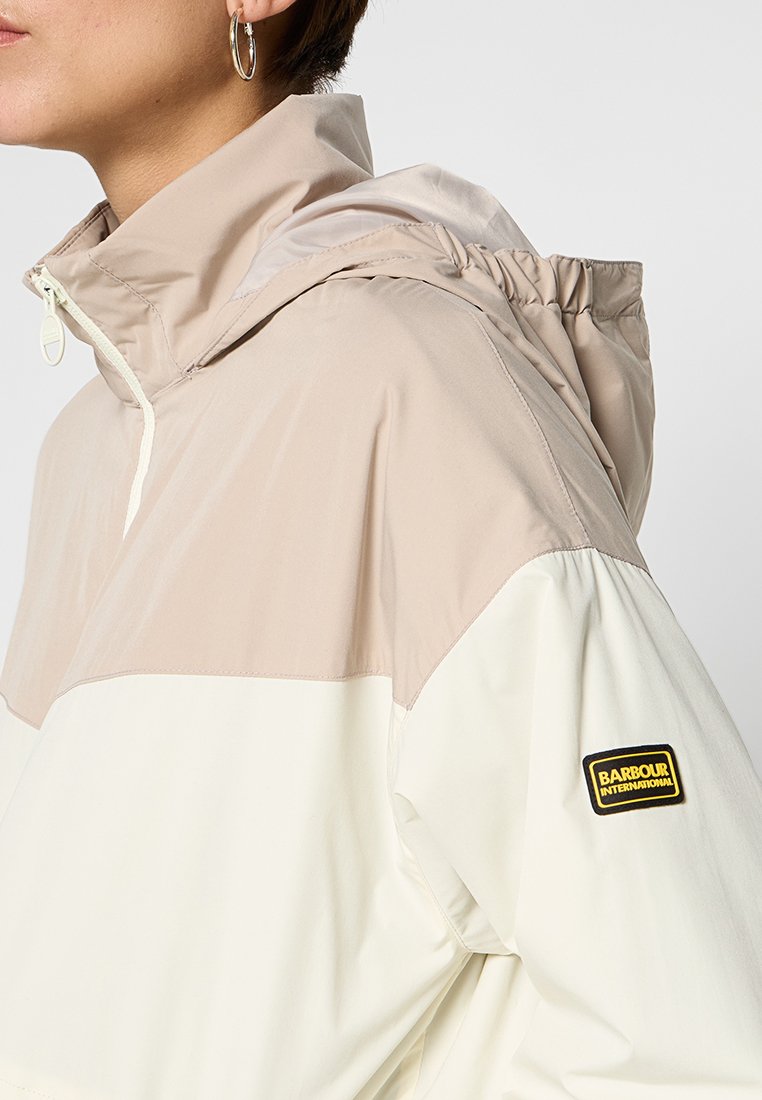 Giacca leggera con cappuccio in crema e beige. Presenta polsini elasticizzati, un cappuccio con cordino e un'etichetta con logo nero e giallo sulla manica.