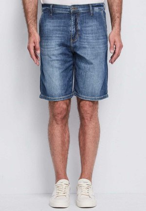 Uomo con pantaloncini di jeans blu fino al ginocchio e scarpe da ginnastica bianche, in piedi davanti a uno sfondo chiaro e uniforme.