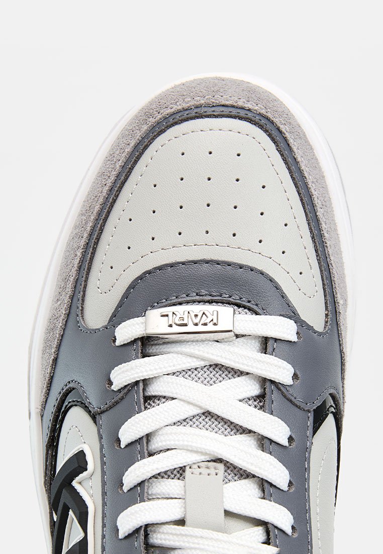Primer plano de la puntera de una zapatilla gris y blanca con cuero perforado, cordones blancos, ribete de ante y etiqueta plateada en los cordones con la inscripción "KARL".