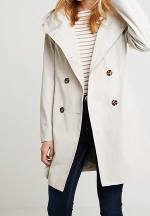 Kobieta ubrana w jasnobeżowy płaszcz dwurzędowy z dużymi guzikami, nałożony na pasiastą koszulę i ciemne obcisłe dżinsy.