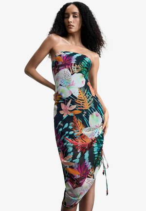 Vrouw met krullend haar, gekleed in een strapless, aansluitende midi-jurk met een kleurrijk tropisch bloemenpatroon, poserend met de handen in de zij.