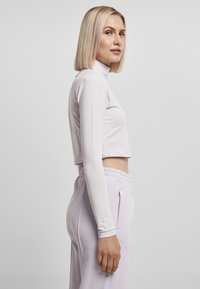 Hvit langermet croppet topp med høy hals, sammen med lyse lilla, høyt liv joggebukser med sidelommer. Glatt stofftekstur.