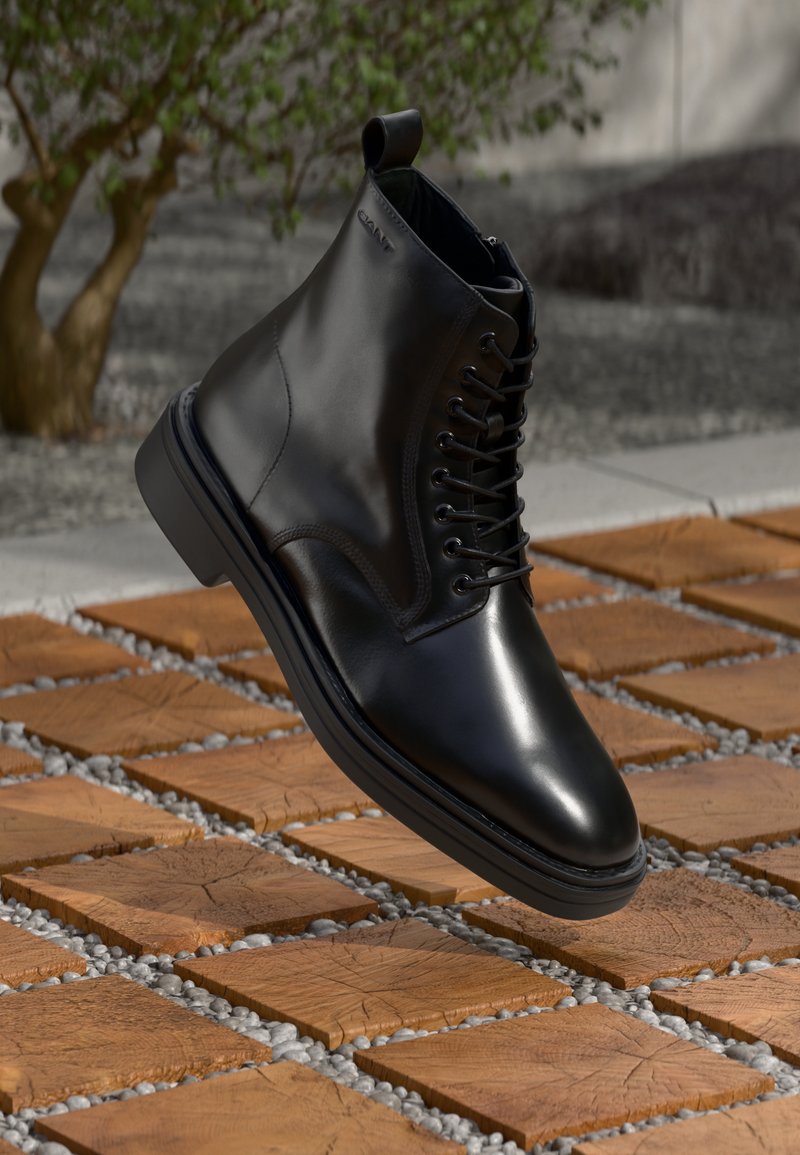 Black leather ankle boot with a smooth finish, lace-up design, and a raised back loop, hovering above a patterned wooden surface.