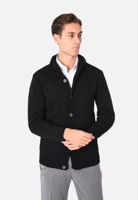 Cardigan nero lavorato a maglia con chiusura a bottoni, polsini e orlo a coste, dotato di due tasche frontali e colletto a scialle.
