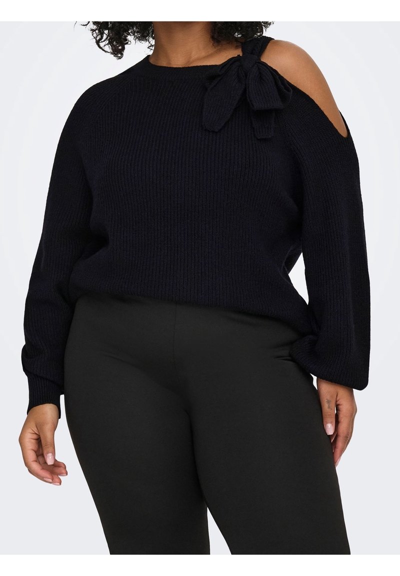 Femme portant un pull noir en maille avec un détail nœud et une découpe épaule dénudée, associée à un pantalon noir ajusté.