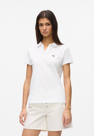 Kvinde iført en hvid kortærmet polo med et lille broderet logo og off-white shorts med knapper, stående mod en ensfarvet baggrund.