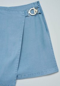 Minifalda de mezclilla azul claro con diseño en envoltura, cremallera lateral y un detalle de hebilla circular metálica para el cierre. Textura suave.