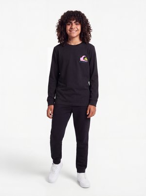 Enfant souriant aux cheveux bouclés, portant un t-shirt noir à manches longues avec un logo coloré, un pantalon noir assorti et des baskets blanches sur fond blanc.